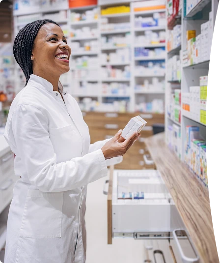 A pharmacist smiling while reviewing shelved medication in a pharmacy.