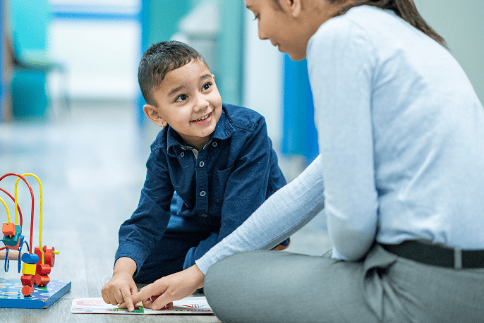 A woman sits on the floor, smiling at a young boy who is also sitting and smiling at her. He is pointing at something in a book lying on the floor between them, next to a bead maze toy.
